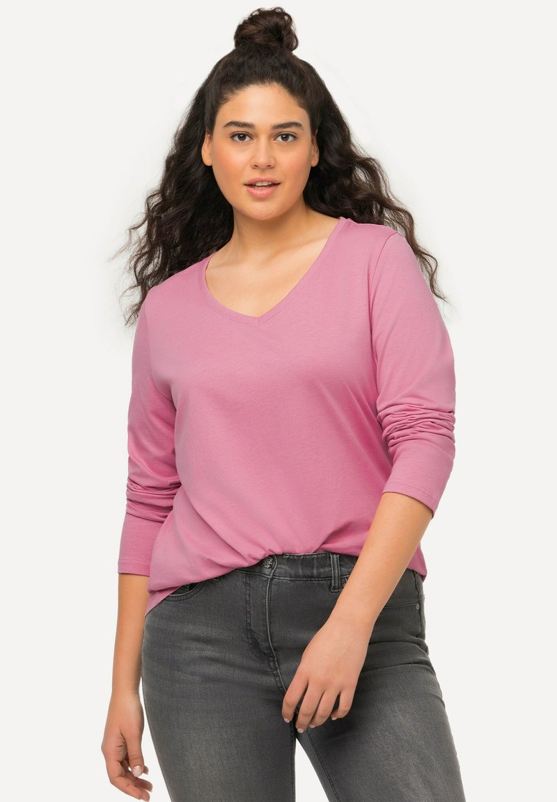 Long-sleeve pink top with a V-neck and relaxed fit, featuring a knotted detail at the waist. Paired with dark gray jeans.