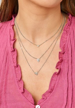 Tres collares plateados de capas con colgantes de corazón y pájaro, llevados en el cuello sobre una blusa rosa con volantes.