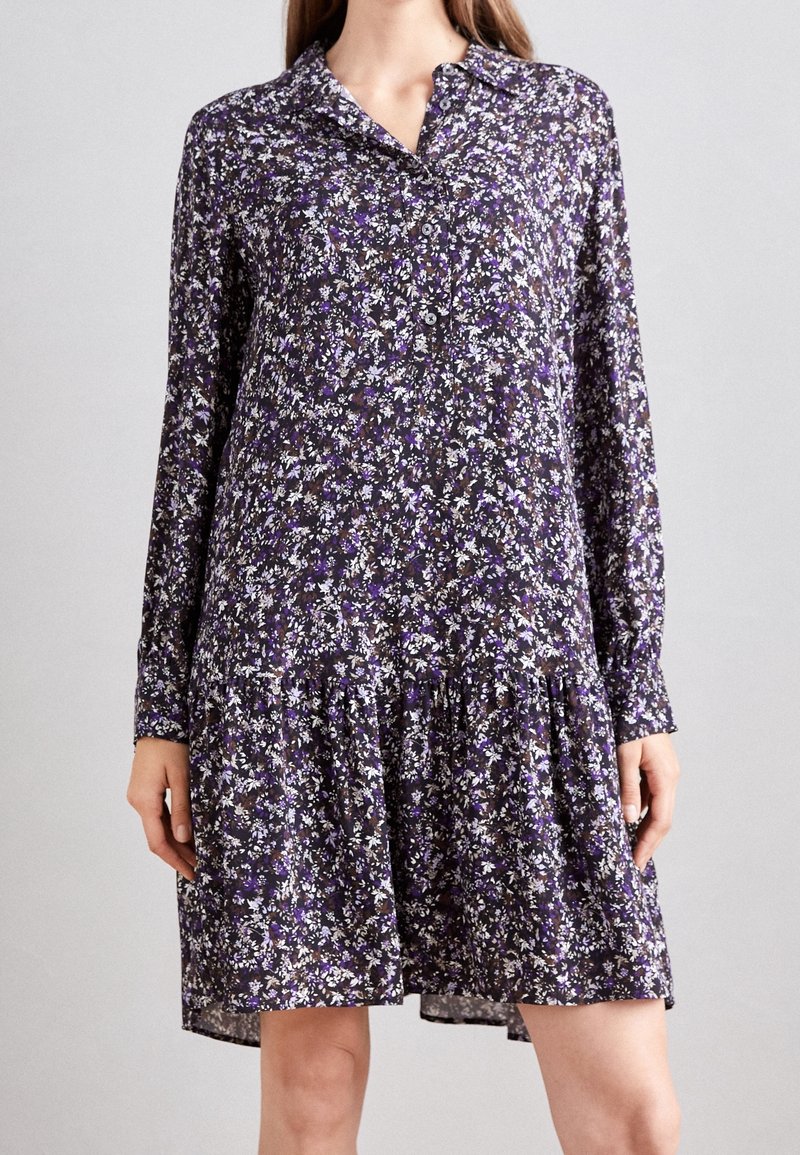 Femme portant une robe ample, à manches longues, longueur genou, à imprimé floral violet et blanc, debout devant un fond uni.