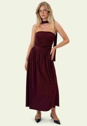Femme blonde portant une robe longue bustier rouge foncé avec une écharpe assortie, des talons noirs pointus et des bijoux minimalistes, debout devant un fond uni.