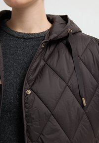 Bruine gewatteerde jas met capuchon, voorzien van gouden hardware, een gestructureerde stof en een diagonaal ruitpatroon, gelaagd over een donkere trui.