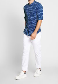 Blå skjorta med blommönster, långa ärmar uppkavlade, kombinerad med vita byxor och vita sneakers med grå detaljer.