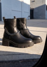 Ζεύγος μαύρων δερμάτινων μποτών αστραγάλου με χοντρές σόλες και ασημί αλυσίδα, τοποθετημένα σε ηλιόλουστο αστικό πεζοδρόμιο.