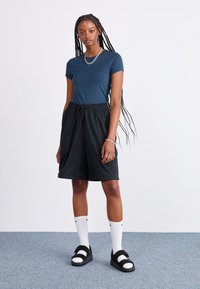 Sieviete ar garām pīņām, kas valkā zilu t-kreklu, melnas šortus ar izvelkamu auklu, baltas Nike zeķes, melnus čības un sudraba ķēžu rotaslietas, stāv uz pelēka paklāja.