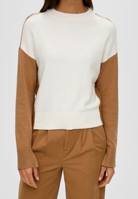 Femme portant un pull à manches longues blanc et beige associé à un pantalon taille haute beige sur un fond uni.