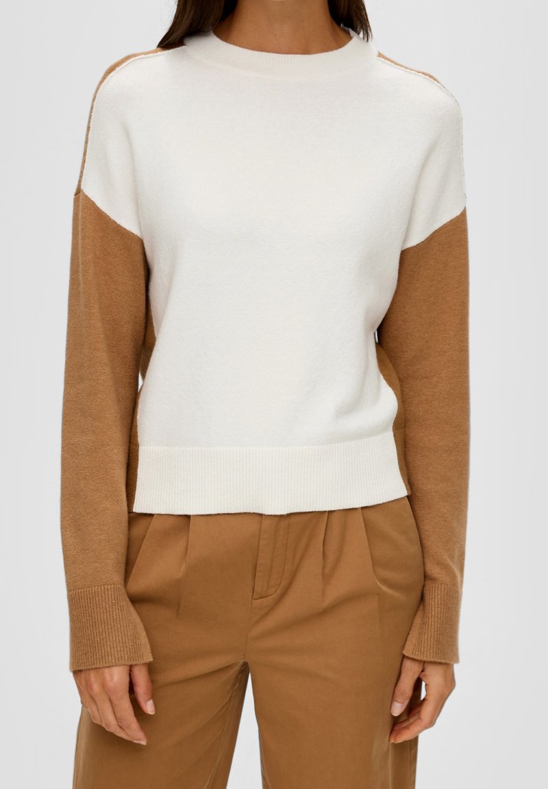 Femme portant un pull à manches longues blanc et beige associé à un pantalon taille haute beige sur un fond uni.