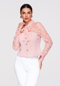 Femme portant un chemisier rose à manches en dentelle et col lavallière, associé à un pantalon blanc, debout devant un fond uni.