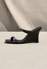 Sandale compensée noire avec deux fines lanières, une sur les orteils et une près de la cheville, sur un fond beige texturé.