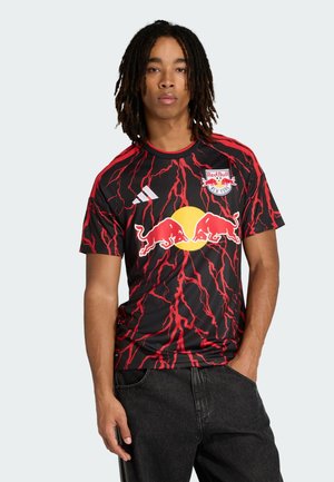 Ung mand med dreadlocks iført en sort og rød New York Red Bulls fodboldtrøje med lynmønster, poserer med den ene hånd i lommen.