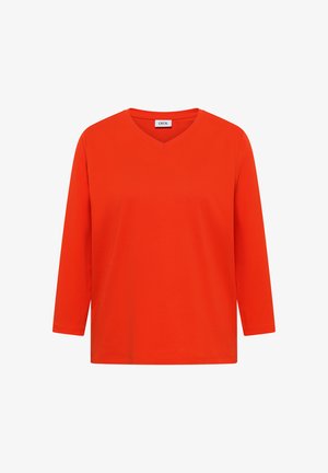 Chemise orange vif à manches longues avec un col en V et une petite étiquette blanche au niveau du col portant l'inscription "CECIL", sur fond blanc uni.