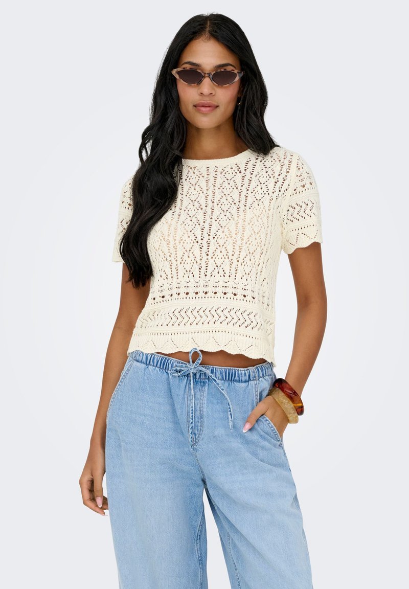 Haut tricoté couleur crème avec un ourlet festonné, manches courtes et un motif complexe rappelant de la dentelle. Associé à un jeans bleu clair fluide.