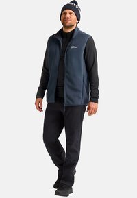 Marineblauer Fleece-Weste mit Reißverschluss über einem schwarzen Langarmshirt, kombiniert mit schwarzen Hosen und einer Mütze mit Bommel oben.