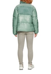 Groene pufferjack met een glanzende afwerking, ribbeltextuur aan de achterkant, gecombineerd met lichtgrijze jeans en witte sneakers.