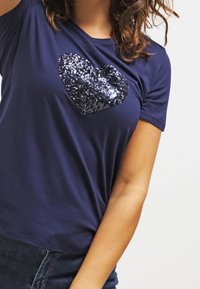Navy blue T-shirt in soft fabric featuring a large, sequined heart design. Short sleeves and a relaxed fit.