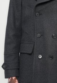 Manteau en laine gris avec un design à double boutonnage, de grands boutons noirs, des revers prononcés et une finition texturée avec une poche latérale.