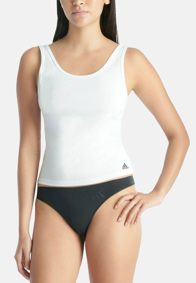 Femme portant un débardeur blanc côtelé sans manches et une culotte de bikini noire, debout avec une main sur la hanche.