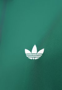 Grønt tekstil med en brodert hvit logo med tre horisontale striper og stiliserte blader, teksturert overflate med en glatt finish.