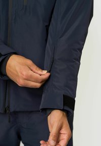 Person, der den Reißverschluss des Ärmels einer dunkelblauen Jacke mit einem Klettverschluss am Handgelenk anpasst, vor einem schlichten hellen Hintergrund.