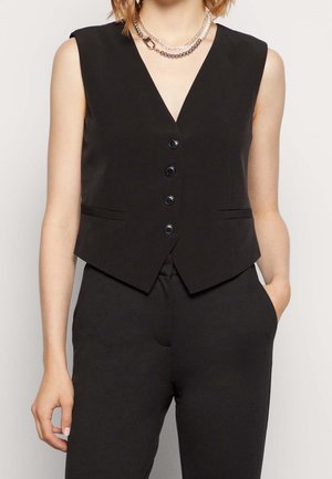 Vrouw die een zwarte, mouwloze V-hals gilet met knopen draagt en bijpassende broek, gecombineerd met meerdere zilveren kettingen.