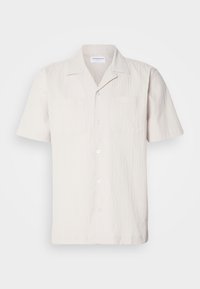 Camisa de manga corta color beige claro con un tejido texturizado, cuello con muesca y dos bolsillos en el pecho, presenta un cierre frontal con botones.