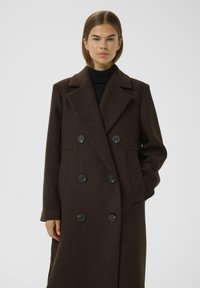 Manteau en laine brun avec un design croisé, grands revers et boutons noirs. Dispose de poches latérales et d'une coupe décontractée.