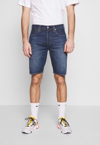 Shorts de mezclilla en azul oscuro, de largo hasta el medio muslo, con cinco bolsillos y detalles de costura. Se llevan con zapatillas blancas y calcetines cortos.