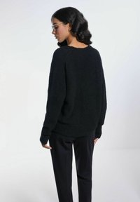 Czarny, puszysty sweter z okrągłym dekoltem, opuszczonymi ramionami i luźnym krojem, zestawiony z czarnymi spodniami. Widok z tyłu.