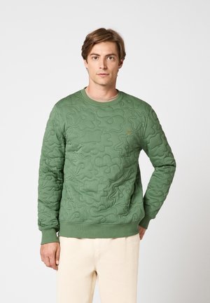 Homme portant un sweat-shirt matelassé vert avec des motifs floraux abstraits et un pantalon beige, debout devant un fond clair uni.