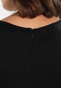 Haut noir avec un large décolleté, un tissu lisse et une fermeture éclair discrète à l'arrière. Design simple et élégant.