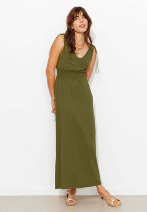 Femme portant une robe maxi sans manches vert olive avec taille froncée et bretelles nouées, debout contre un mur blanc, portant des sandales et des bijoux dorés.