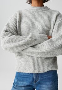 Person, der einen hellgrauen gerippten Strickpullover mit langen Ärmeln trägt, die Arme verschränkt, sowie blaue Jeans vor einem schlichten Hintergrund.