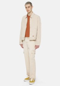 Giacca di jeans beige, maglietta arancione e pantaloni cargo beige. Sneakers con dettagli gialli. Design semplice, taglio dritto e dettagli pratici.