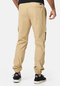 Pantalones cargo beige con puños elásticos, que cuentan con bolsillos laterales y un bolsillo trasero tipo parche, fabricados con una tela ligera y suave. Zapatillas negras.