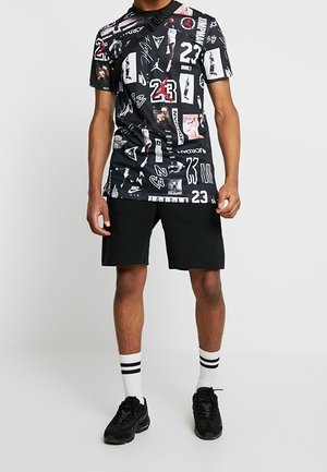 Schwarzes Graphic-T-Shirt mit unterschiedlichen Designs, Logos und Zahlen, kombiniert mit schwarzen Shorts und schwarzen Sneakers. Weiße Socken mit schwarzen Streifen.