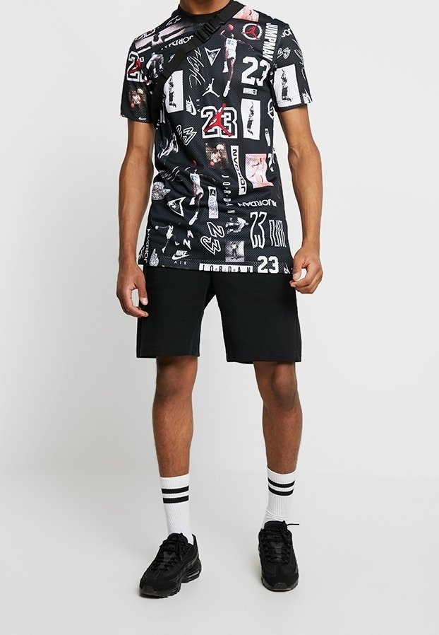 Camiseta gráfica negra con diseños variados, logotipos y números, combinada con pantalones cortos negros y zapatillas negras. Calcetines blancos con rayas negras.
