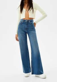 Valkoinen ribbattu crop top, jossa syvä V-pääntie, yhdistetty korkeavyötäröisiin leveälahkeisiin sinisiin farkkuihin ja valkoisiin tennareihin. Sujuva denim-kudos ja saumat.