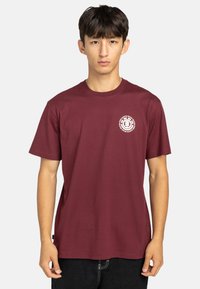 Camiseta de algodón color marrón con mangas cortas y cuello redondo. Presenta un logo blanco circular en el lado izquierdo del pecho. Textura suave, corte relajado.
