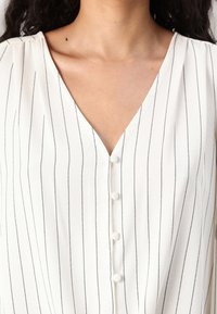 V-neck blouse in a lightweight, cream fabric with vertical black stripes, featuring three buttons and subtle pleating at the shoulders.