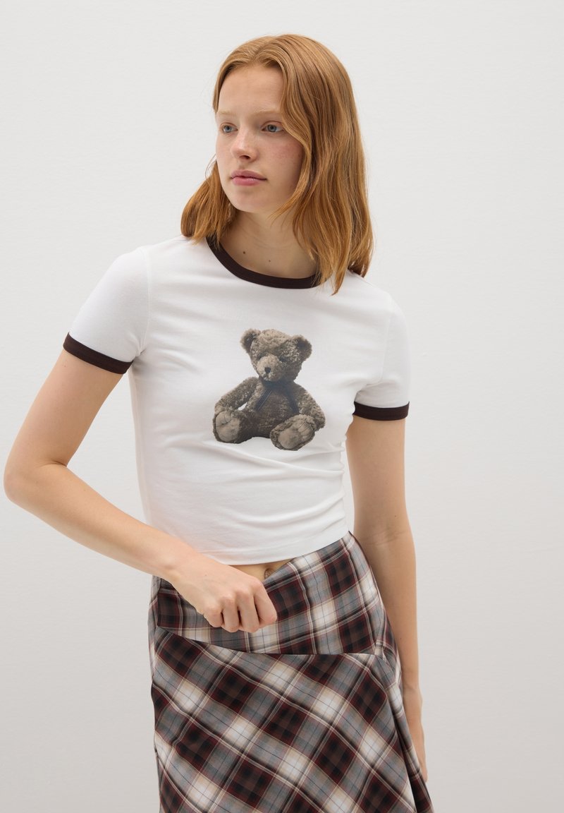 Camiseta de algodón blanca con cuello acanalado marrón que presenta un gráfico de un oso de peluche, combinada con una falda de cuadros en tonos de gris, rojo y crema.