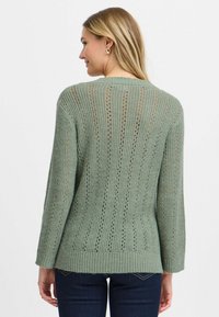 Ljusgrön stickad tröja med ett texturerat, öppet stickmönster, långa ärmar och ribbad nederkant, sedd bakifrån.