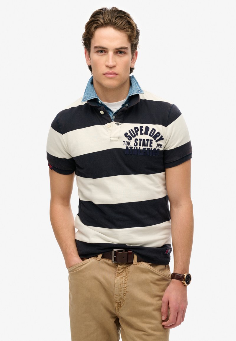 Marineblauwe en crèmekleurige gestreepte poloshirt met een denim kraag, gecombineerd met kaki broek en een bruine riem. Voorzien van een logo-opdruk op de borst.