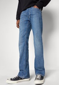 O persoană îmbrăcată în blugi drepți de culoare albastră și adidași negri cu talpă și șireți albi, stând cu o mână în buzunar.