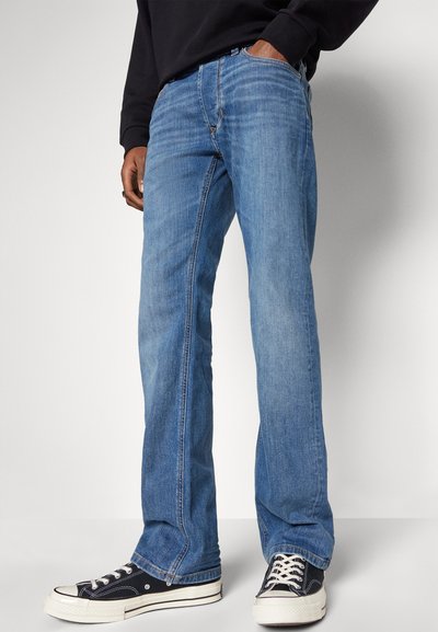 Personne portant un jean droit bleu et des baskets basses noires avec des semelles et des lacets blancs, se tenant avec une main dans la poche.