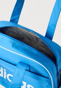 Μπλε αθλητική τσάντα Adidas με λευκό λογότυπο, ανοιχτό φερμουάρ που αποκαλύπτει κενό γκρι εσωτερικό διαμέρισμα και διπλή Schulter ιμάντες.