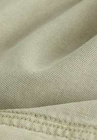Gros plan sur un tissu beige avec un tissage diagonal et une couture double visible, montrant la texture et les plis.