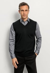 Schwarze ärmellose Strickweste über einem grauen Hemd mit Knöpfen. Die Weste hat ein V-Ausschnitt-Design, eine glatte Stricktextur und eine einfarbige Gestaltung.