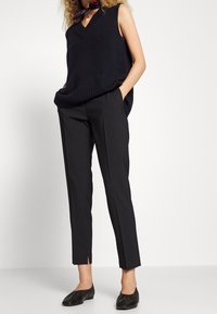 Mörk marinblå ärmlös stickad topp med V-hals och ribbad nederkant, i kombination med skräddarsydda svarta byxor med slitsar i nederkanten och platta svarta skor.