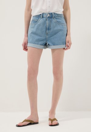 Person trägt hellblaue, hochgekrempelte Jeansshorts, ein weißes ärmelloses Top und braune, flache Sandalen mit Nieten vor einem einfarbigen hellen Hintergrund.