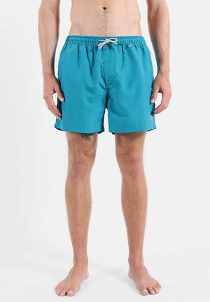 Homme portant un short de bain turquoise avec cordon, debout pieds nus sur fond blanc uni, tatouage visible sur la cuisse gauche.
