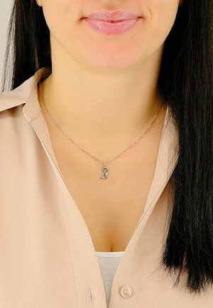 Collana in argento con pendente a forma di piccolo gatto seduto, indossata sopra una maglia a scollo a V, su uno sfondo di tessuto leggero e liscio.